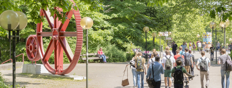 Studenten im Sommer auf dem Nordcampus, rote Zahnräder im Hintergrund